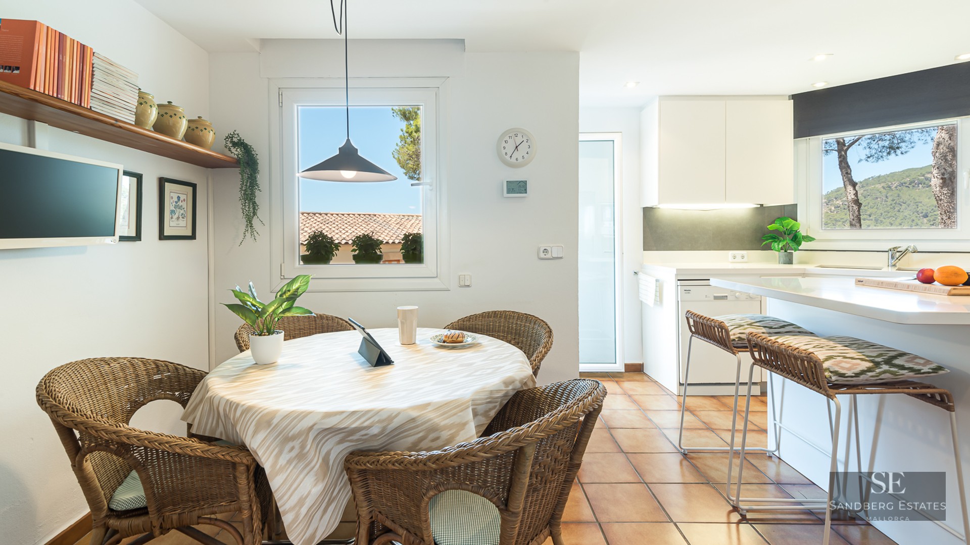 Round dining table with wicker chairs and terracotta flooring next to a modern white kitchen.