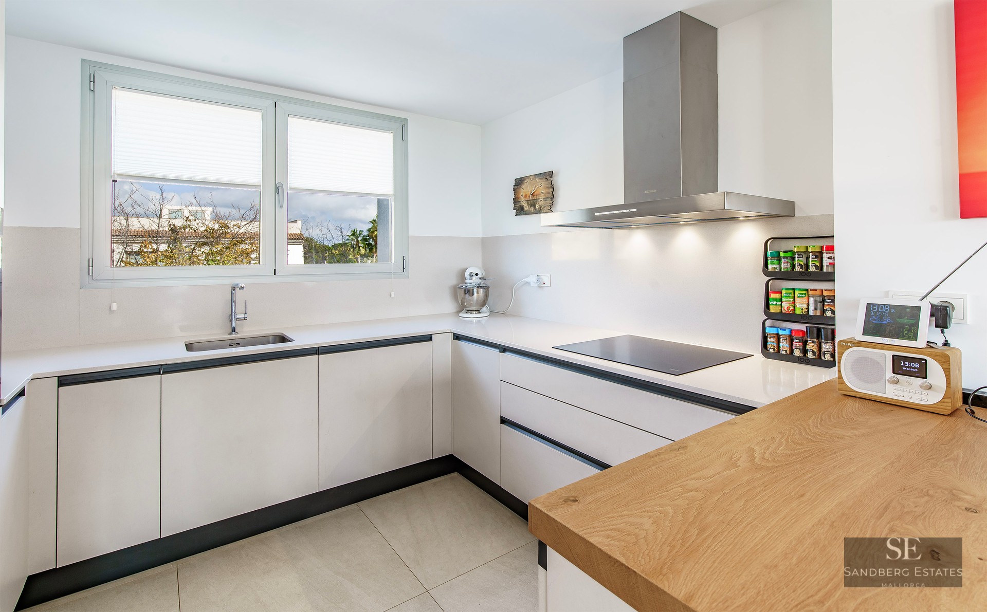 L-shaped modern white kitchen with wood breakfast bar, stainless steel appliances, and a large window.