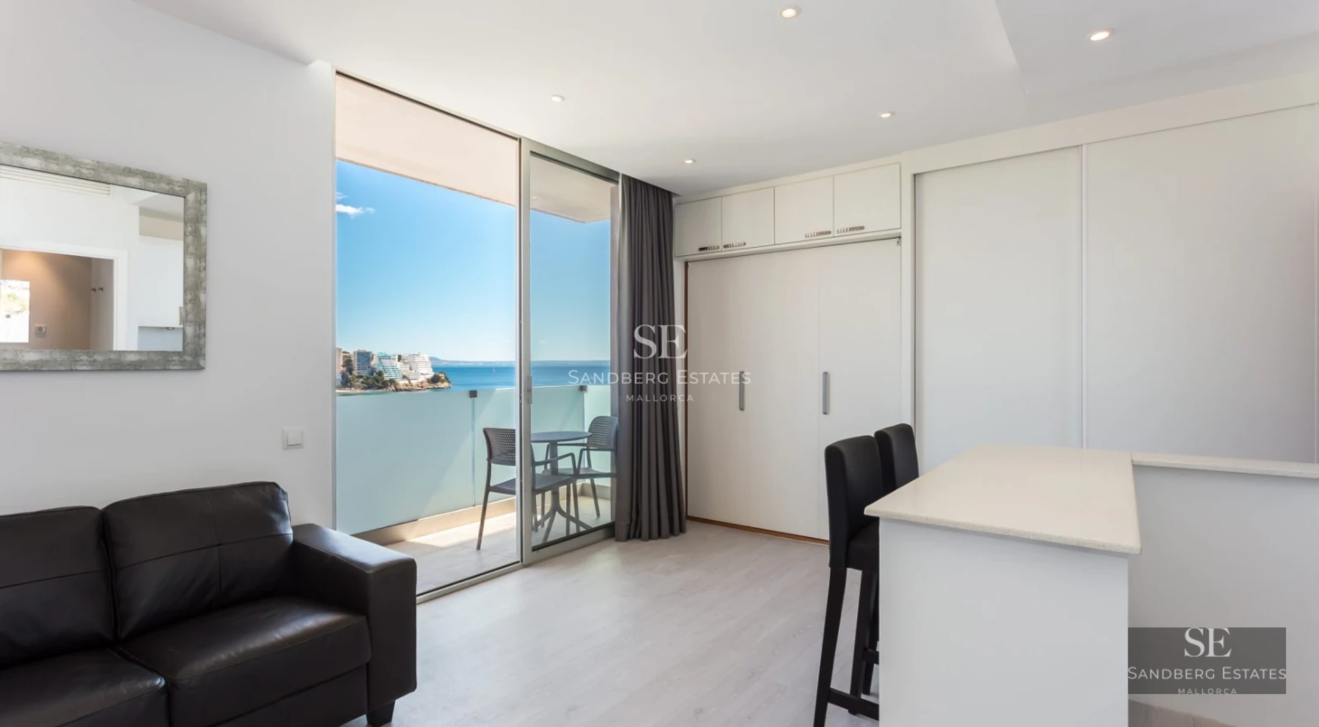 Spacious living room with large windows, light wood floors, and minimalist decor. Natural lighting highlights soft textures and neutral colors.
