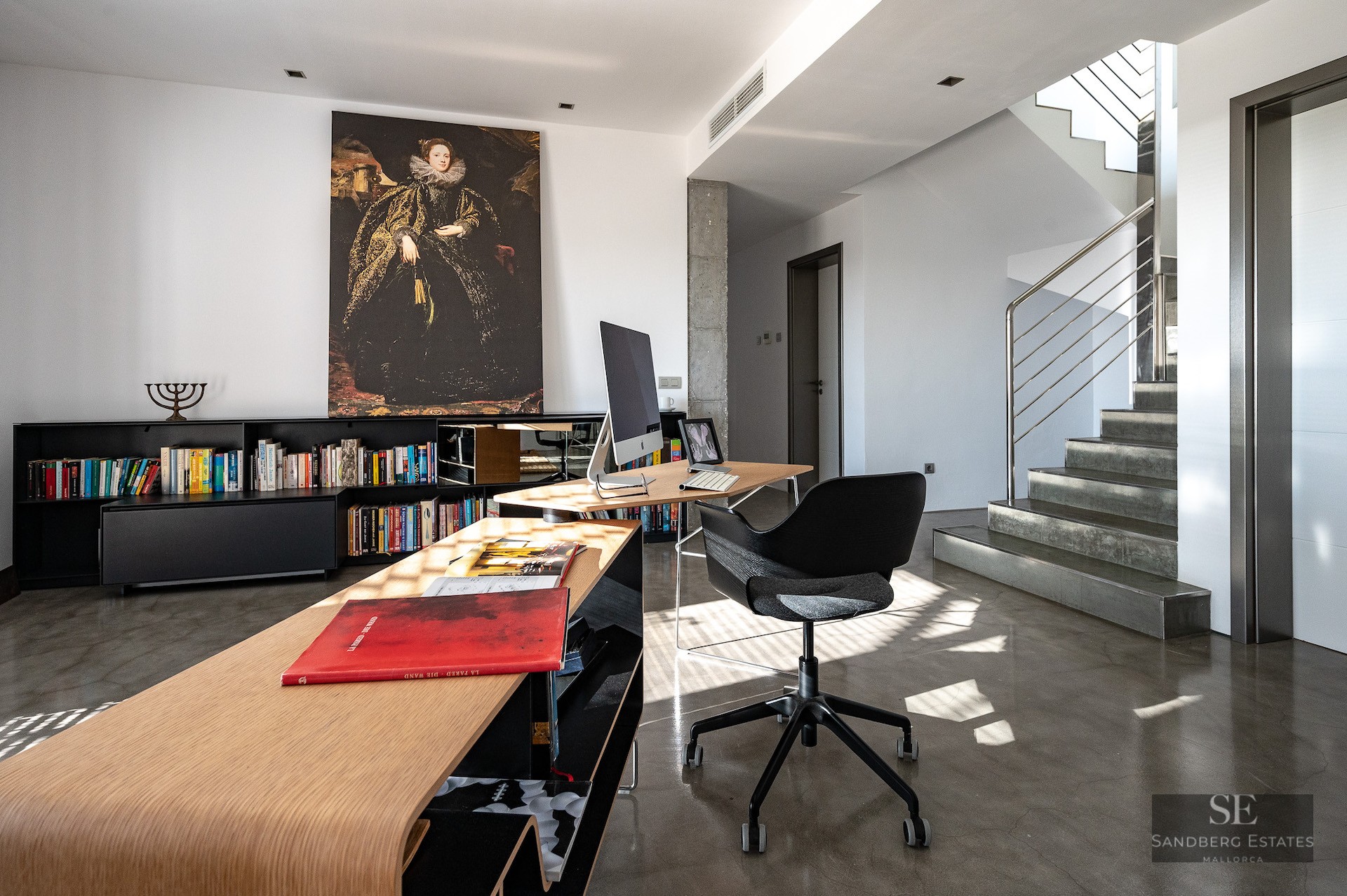 Modern home office with a curved wooden desk, black ergonomic chair, large classical painting, and concrete stairs.