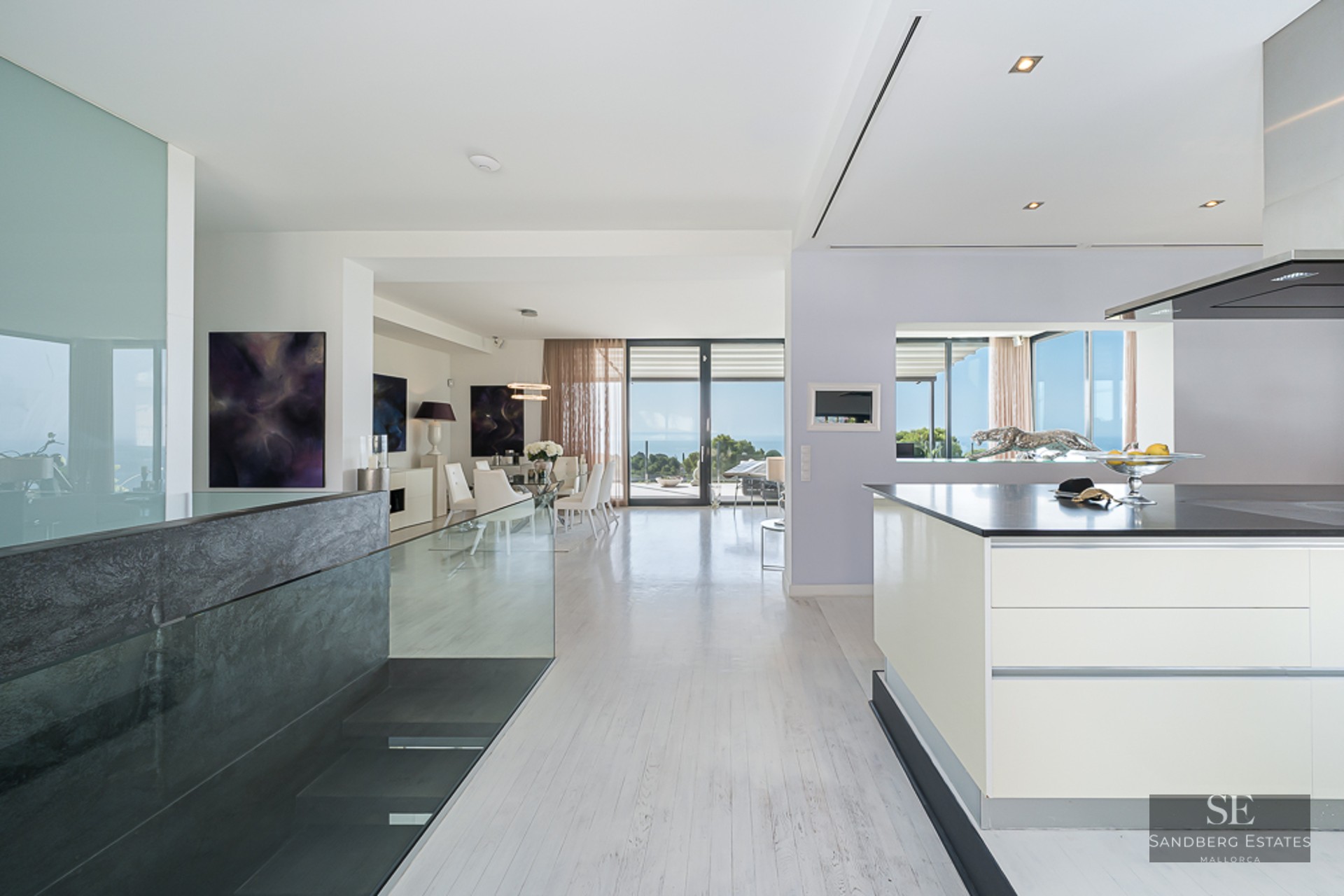 Modern open-plan interior featuring a white kitchen island, glass dining table, and large windows with sea views.