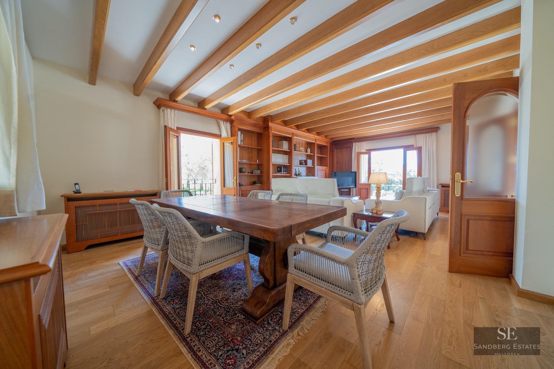 Spacious dining area featuring a solid wood table, wicker chairs, and exposed beam ceiling opening to a living room.
