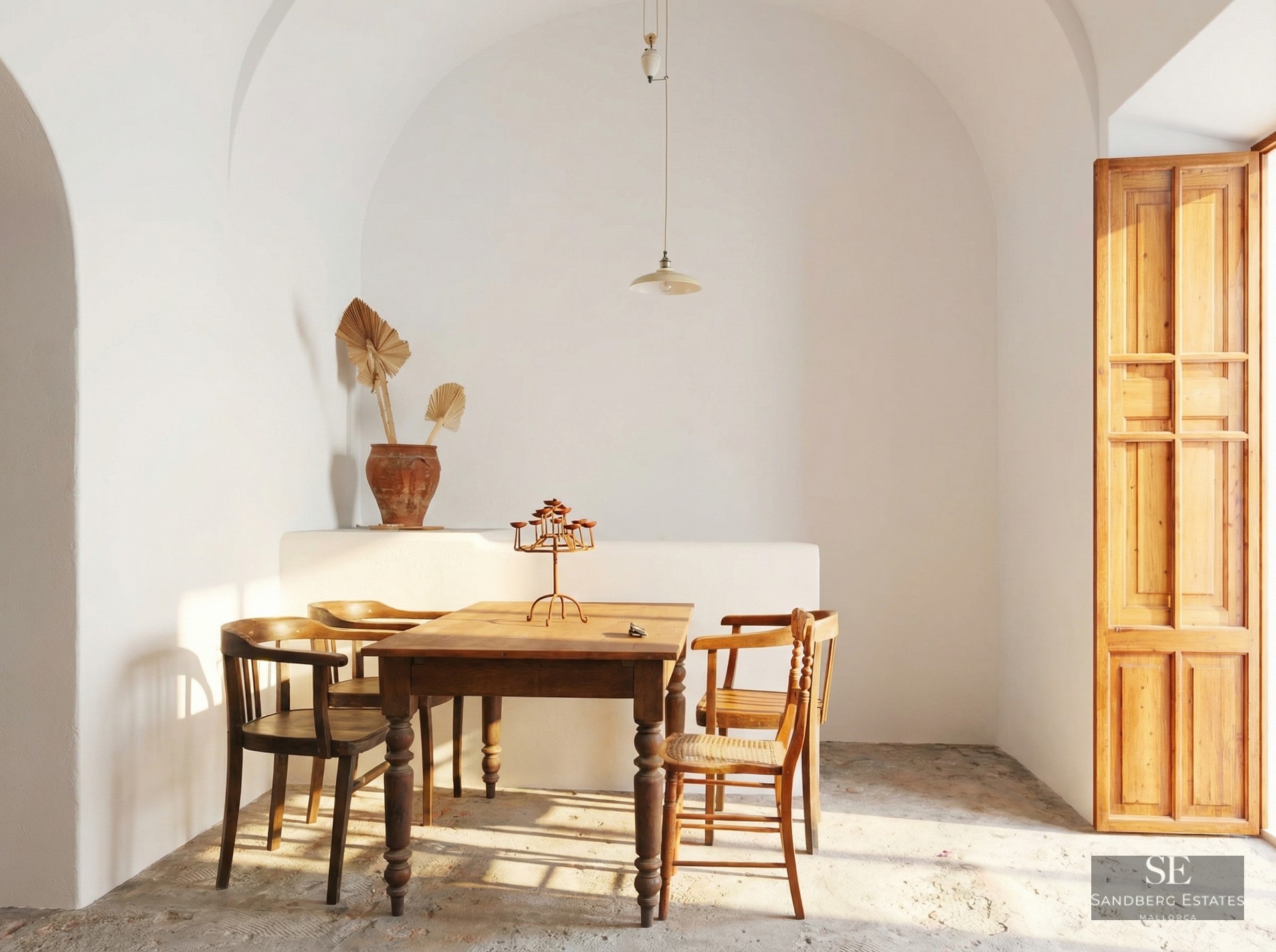 Rustic wooden dining table and four chairs in a white room with a high vaulted ceiling and warm sunlight.