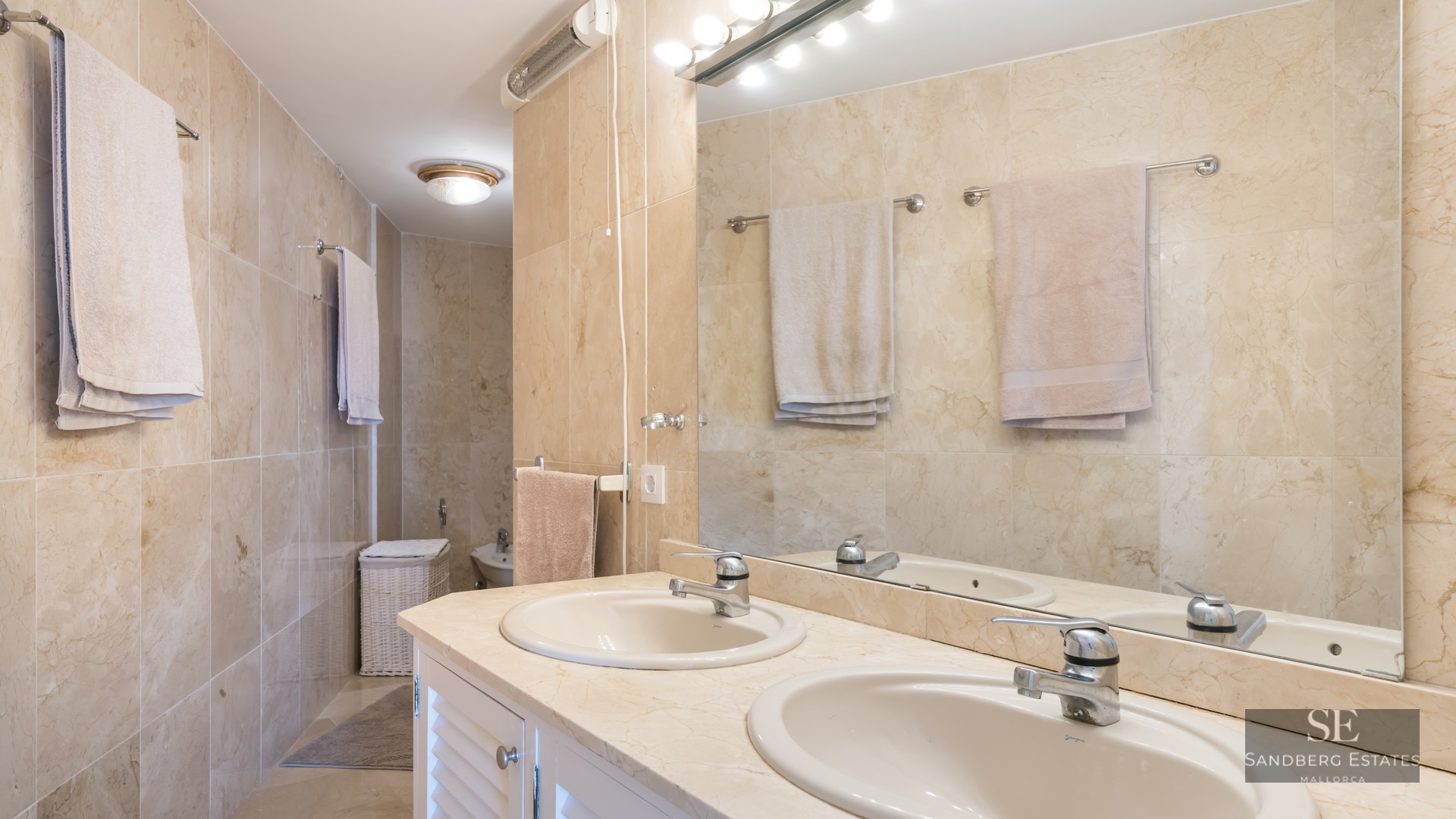 Bathroom featuring a double vanity, beige marble walls, a large mirror, and hanging towels.