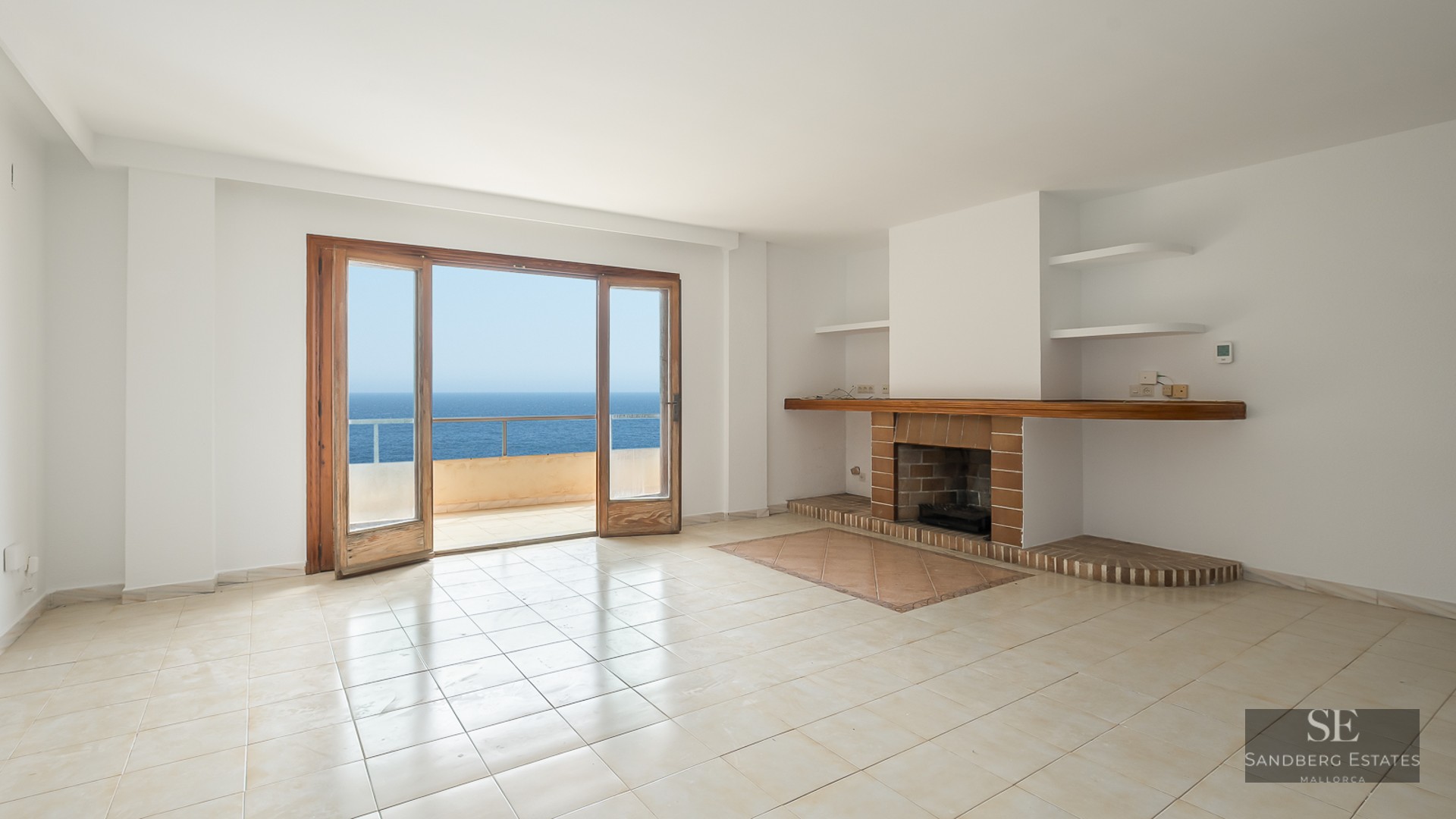 Empty white living room with a brick fireplace and wooden doors opening to a balcony with Mediterranean sea views.