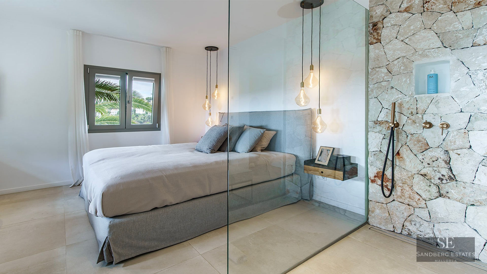 Contemporary bedroom with a grey bed, glass wall divider, and a rustic stone shower with brass fixtures.