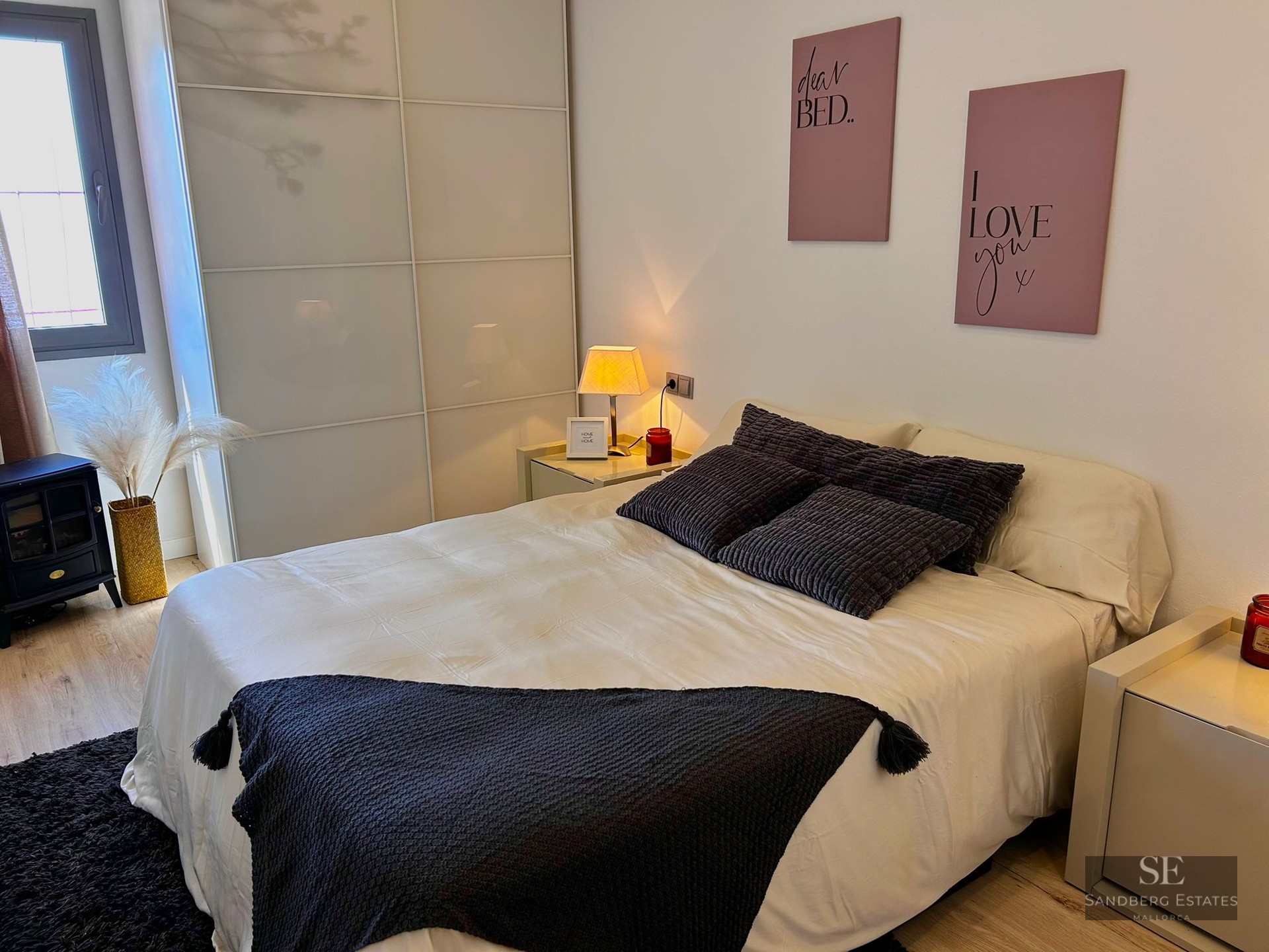 A cozy bedroom with a large bed, dark textured pillows, a frosted glass wardrobe, and soft ambient lighting.