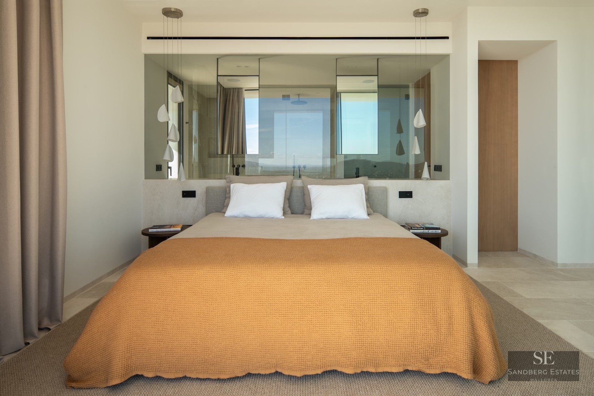 Modern bedroom featuring an orange bedspread, glass partition wall behind the bed, and sculptural white pendant lamps.