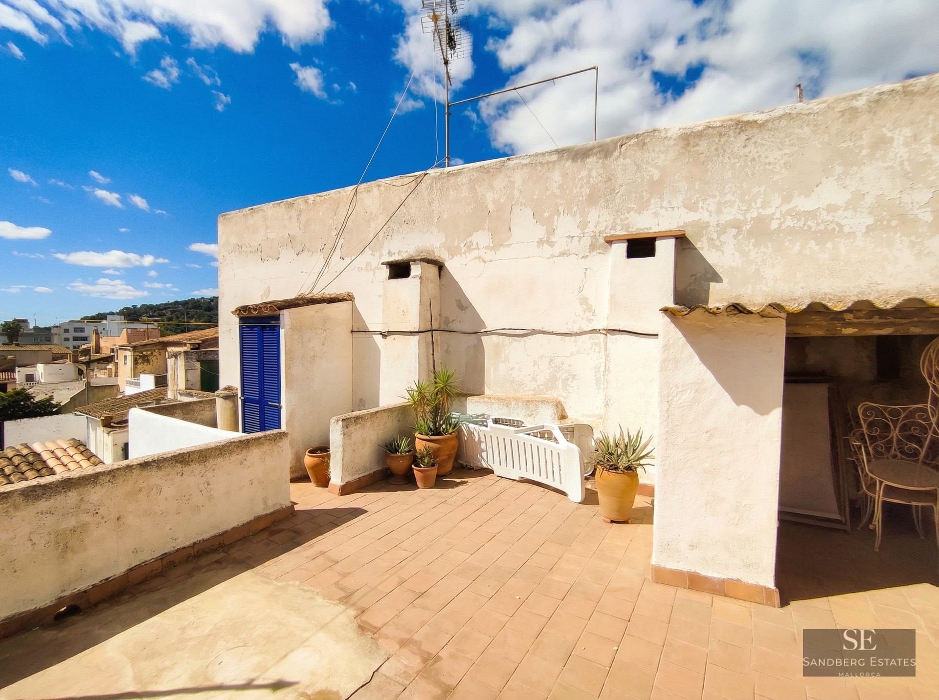 A sunny rooftop terrace with terracotta tiles, white stucco walls, potted plants, and a bright blue door.
