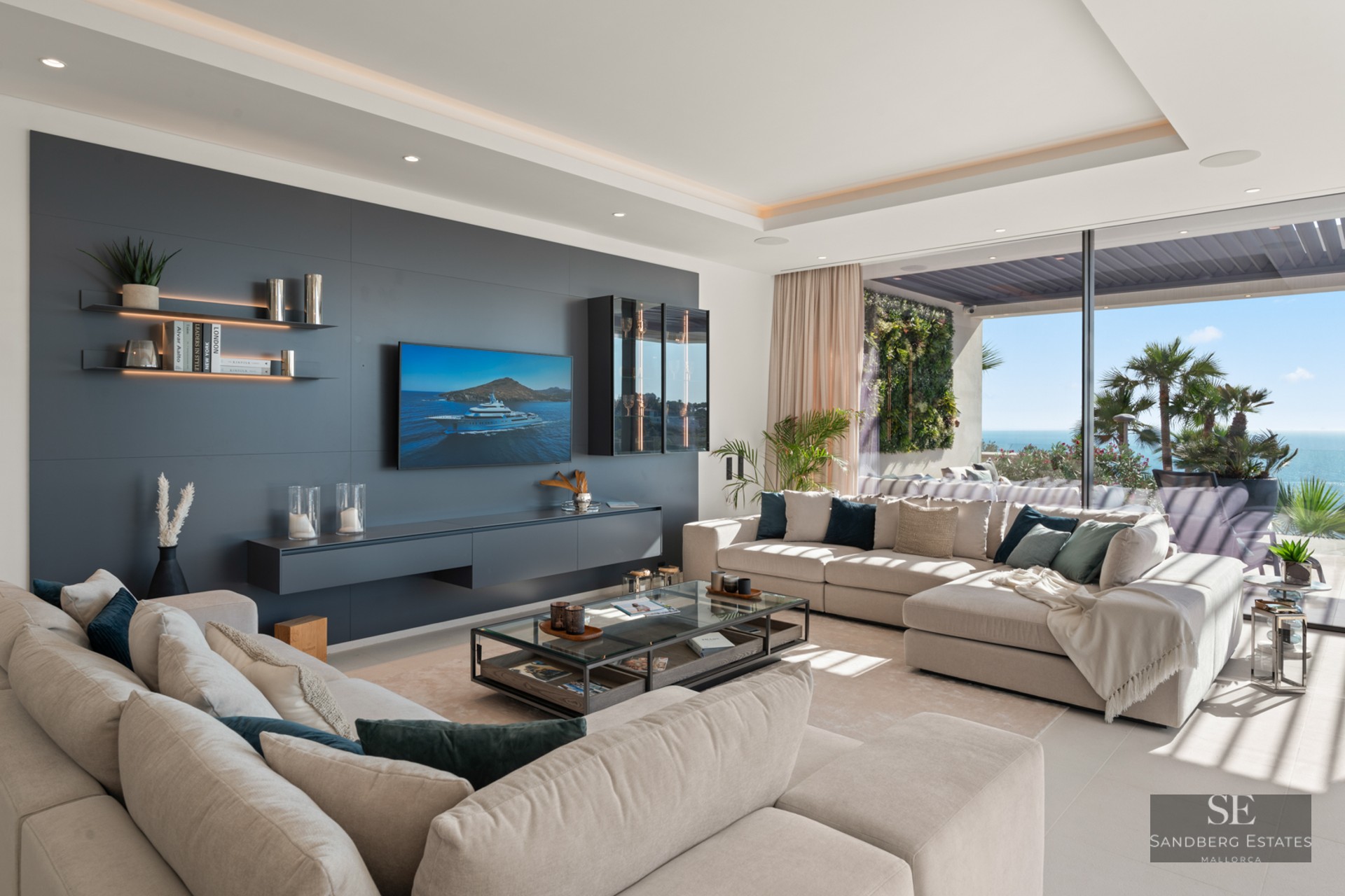 Bright living room with large beige sofas, charcoal accent wall, TV, and panoramic glass doors opening to a sea view terrace.