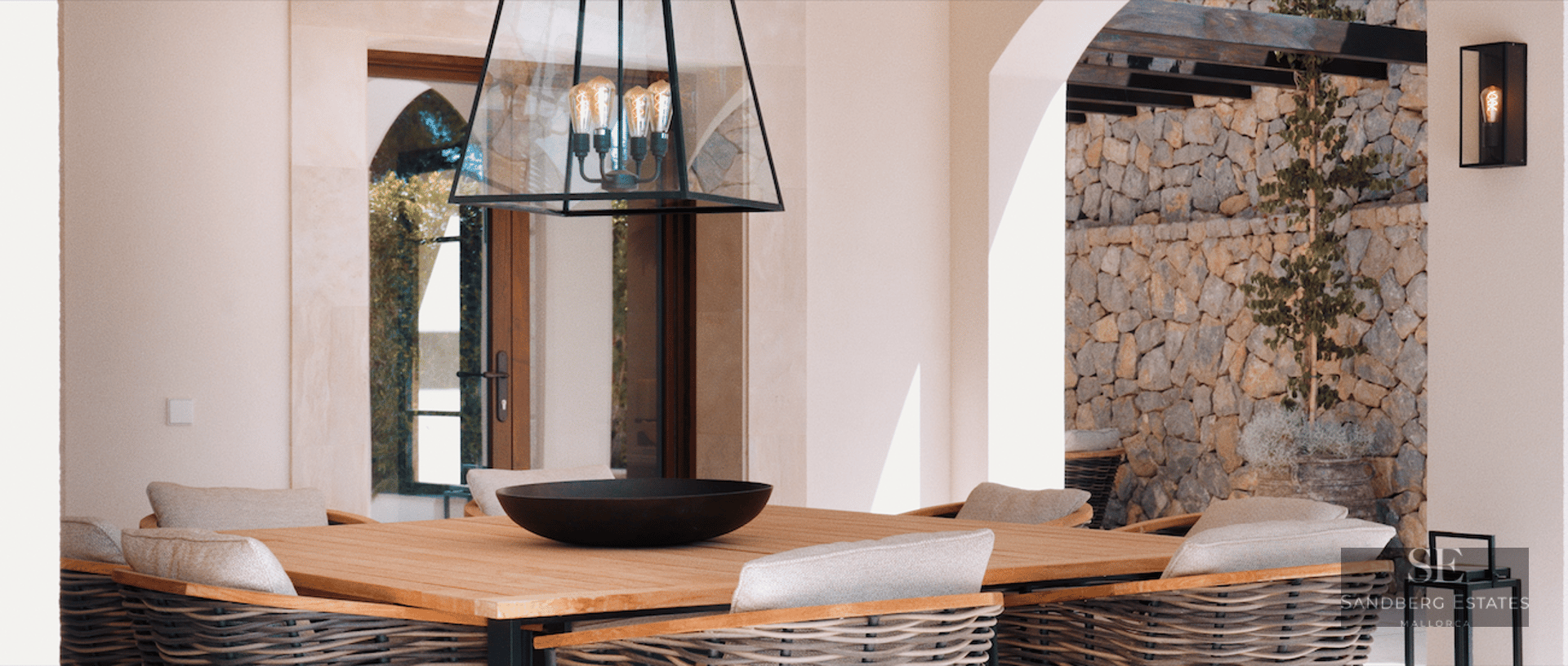 Wooden outdoor dining table with woven chairs on a luxury terrace featuring a stone wall and designer lighting.