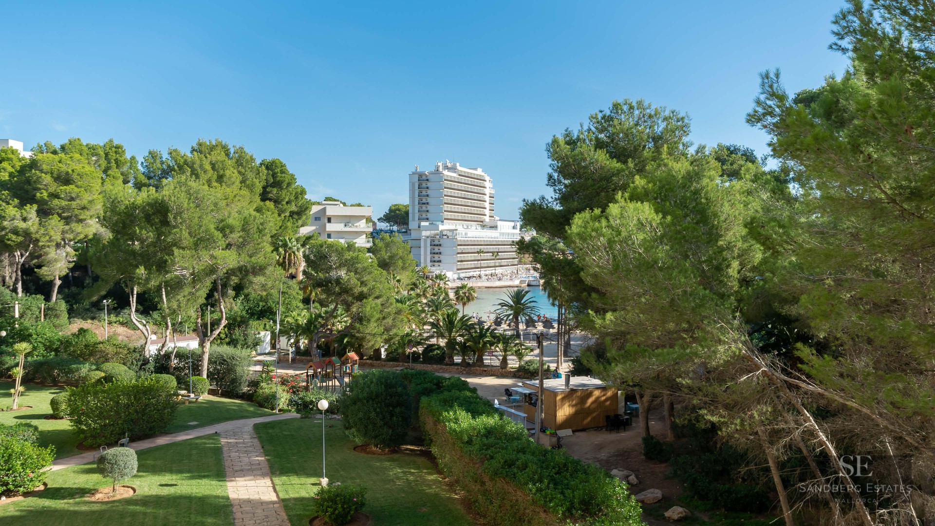 Jardin vert soigné avec des sentiers en pierre, des pins et un grand hôtel surplombant une baie sous un ciel bleu.