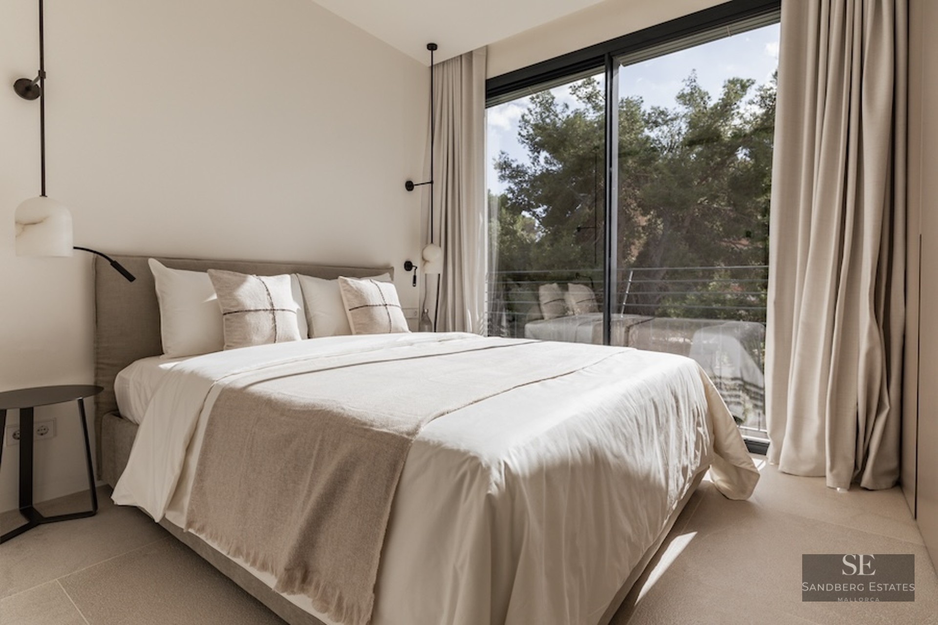 Bright bedroom with neutral-toned bed facing a large floor-to-ceiling window overlooking trees.