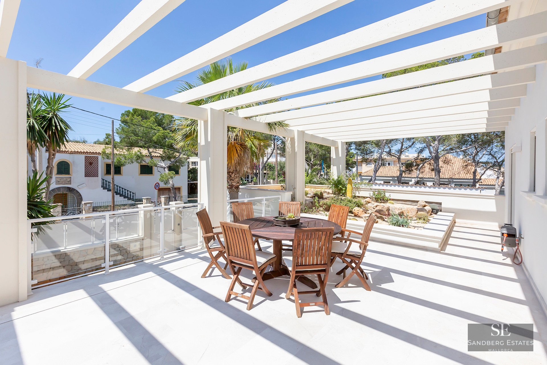 Sunny terrace featuring a white architectural pergola, round wooden dining set, and palm trees under a clear blue sky.