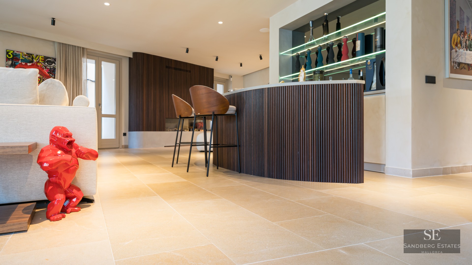 Modern living room featuring a curved wooden bar, designer stools, stone floors, and a bright red gorilla sculpture.