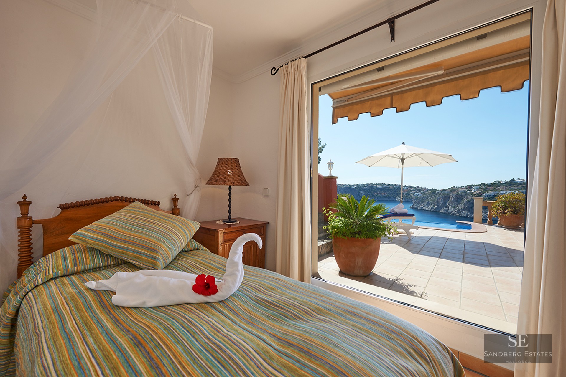 Bedroom with a striped bed and swan towel overlooking a terrace with an infinity pool and blue ocean bay.