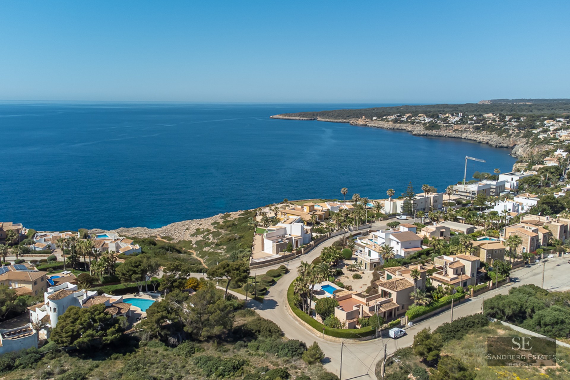 Aerial view of luxury white villas with blue pools along a rocky Mediterranean coastline under a clear blue sky.