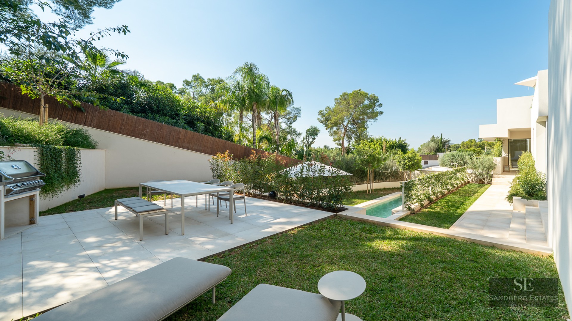 Modern white terrace featuring a dining area, outdoor grill, and lap pool surrounded by lush landscaping and palm trees.