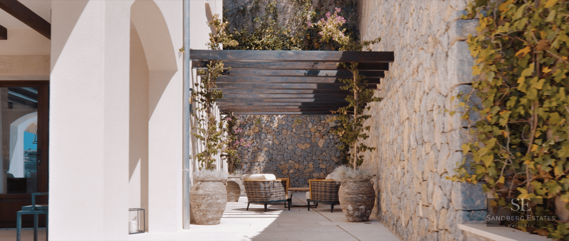 Sunlit Mediterranean terrace featuring rustic stone walls, a dark wood pergola, and comfortable outdoor seating.