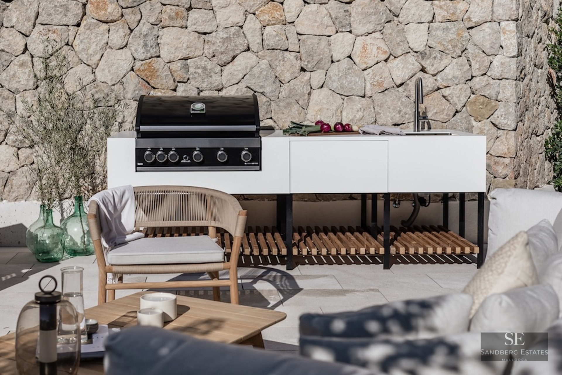 Modern white outdoor kitchen with gas grill and sink against a stone wall with designer patio furniture.