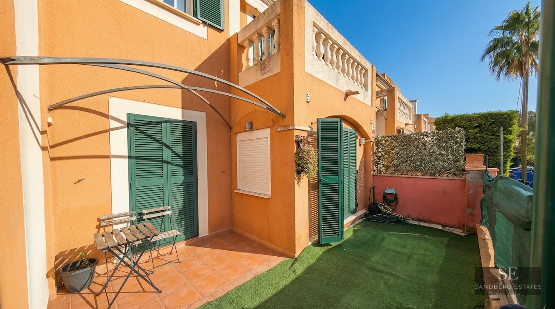 Small private garden with artificial grass, terracotta tiles, green shutters, and a wooden bistro set under a sunny sky.