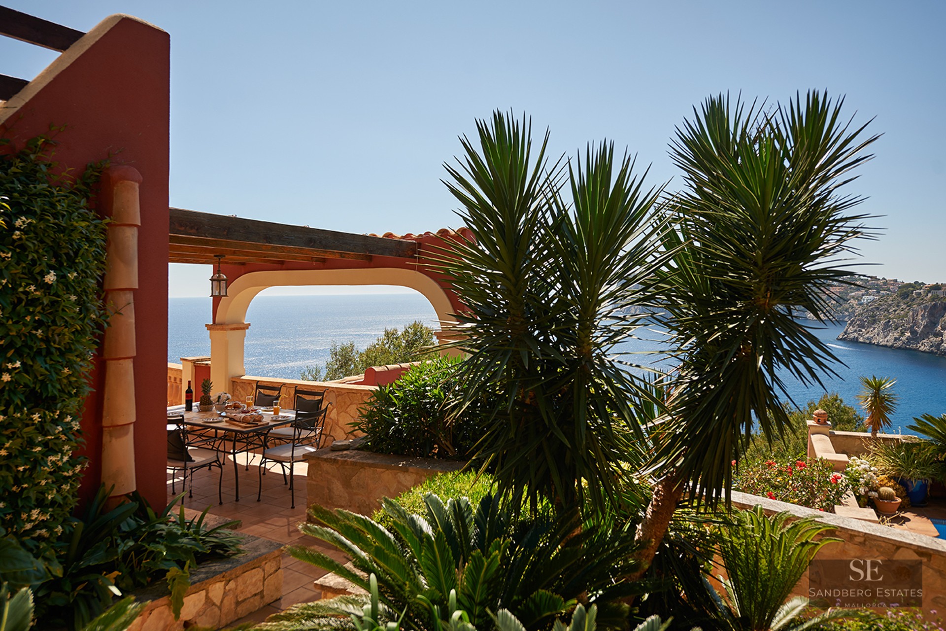 Outdoor dining table on a terracotta terrace with an arched pergola overlooking the blue sea and lush greenery.