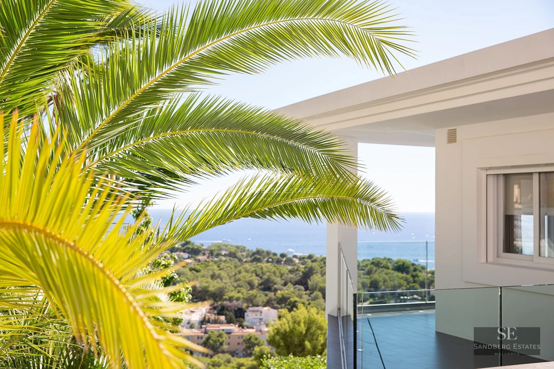 Palm leaves framing a modern white villa terrace with a wide view of the ocean and green hills.