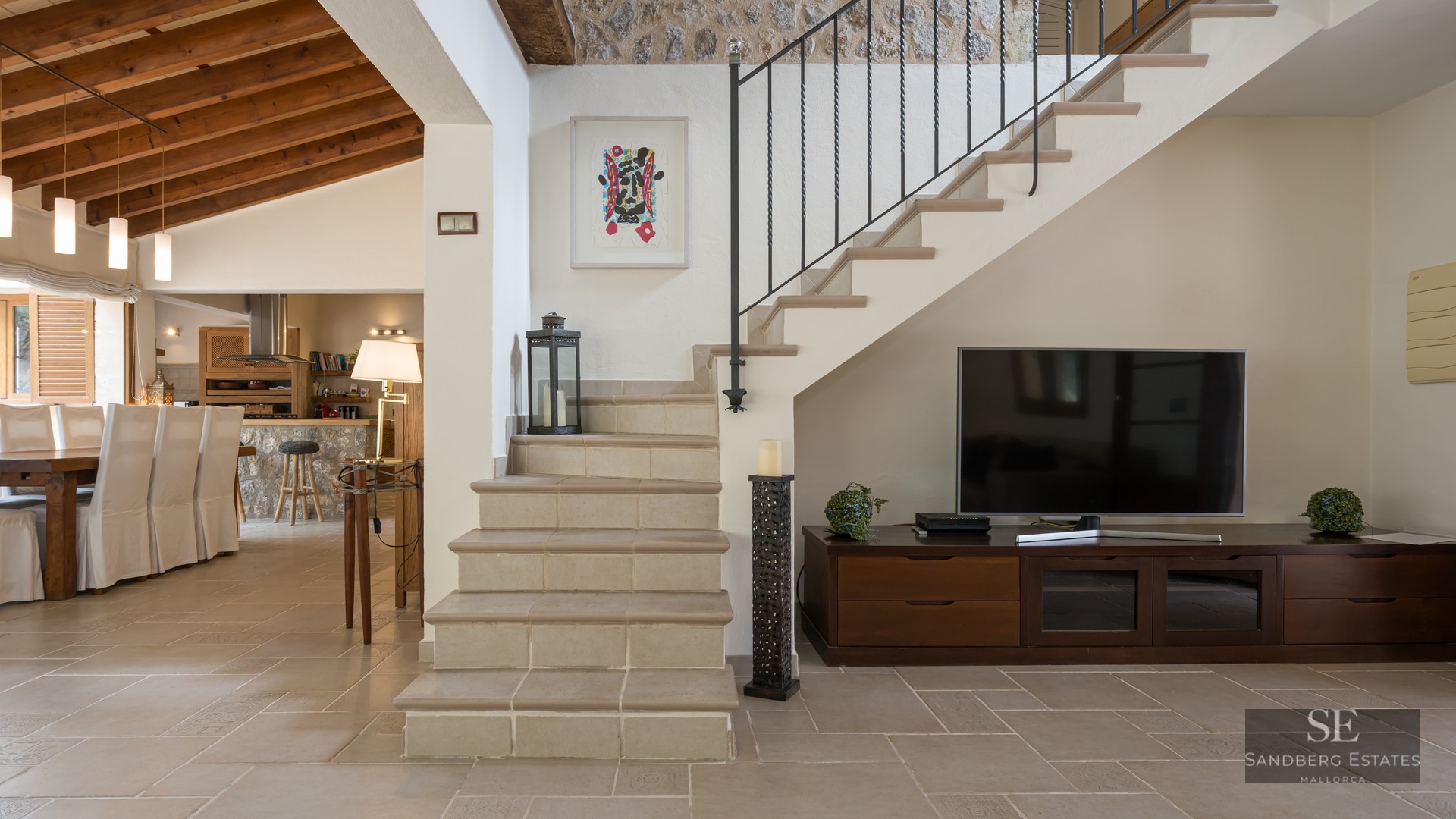 Interior view featuring a stone staircase, wooden TV console, and dining area with exposed ceiling beams.