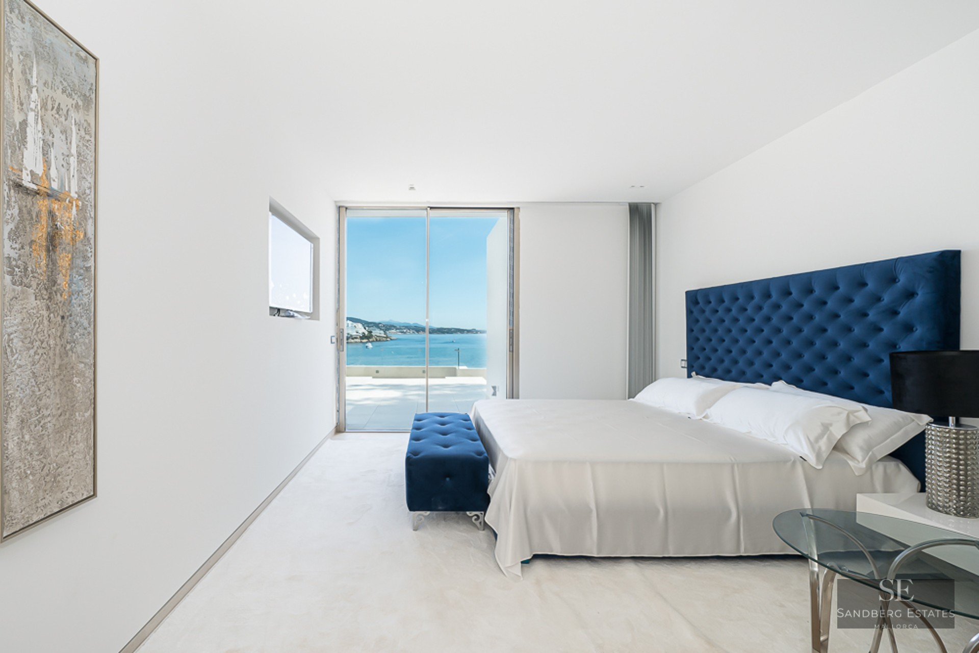 Modern bedroom with blue tufted headboard, white linens, and large glass doors overlooking the ocean.