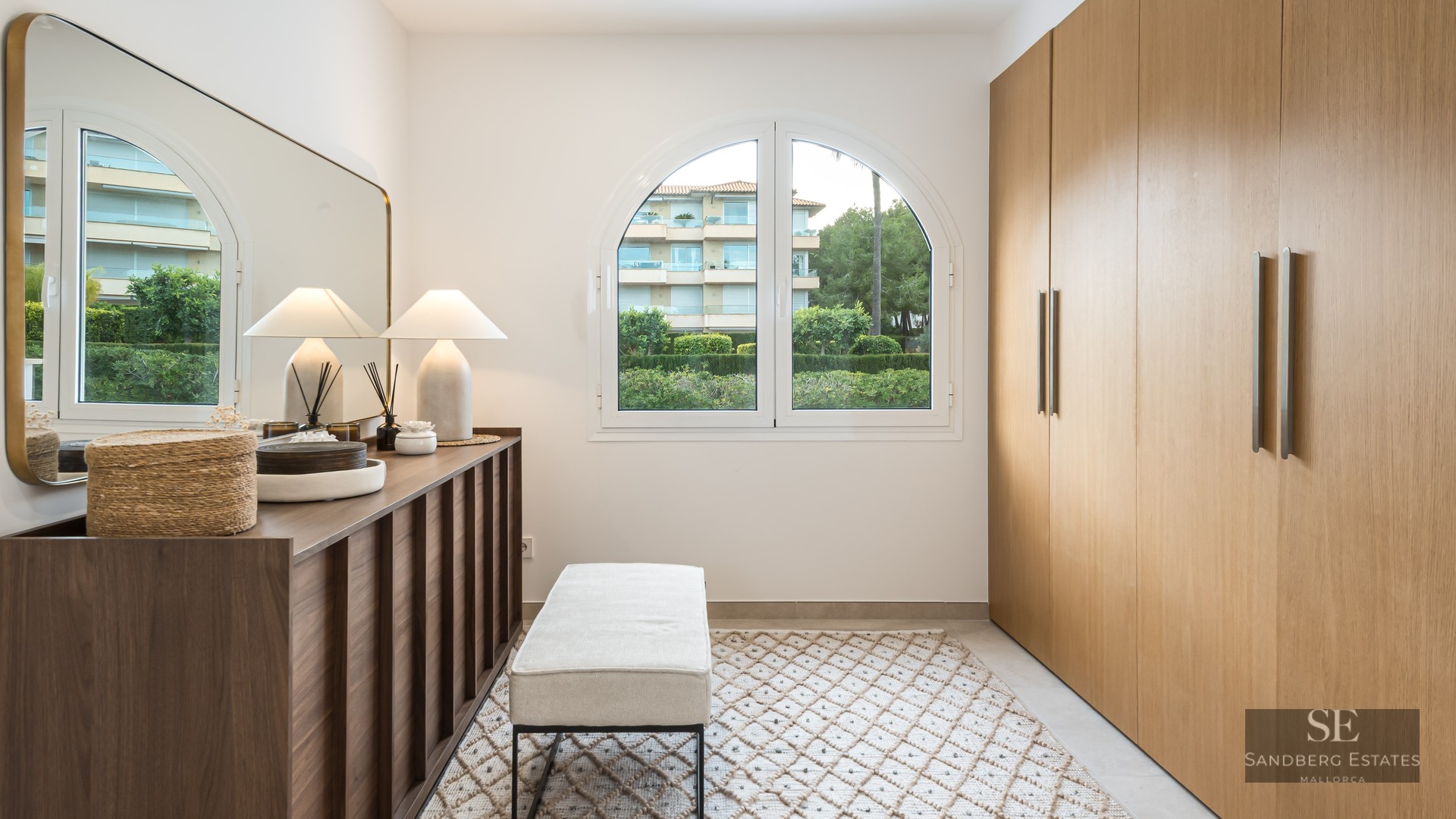 A modern dressing room featuring large oak wardrobes, an arched window, a mirrored console, and a cushioned bench on a rug.