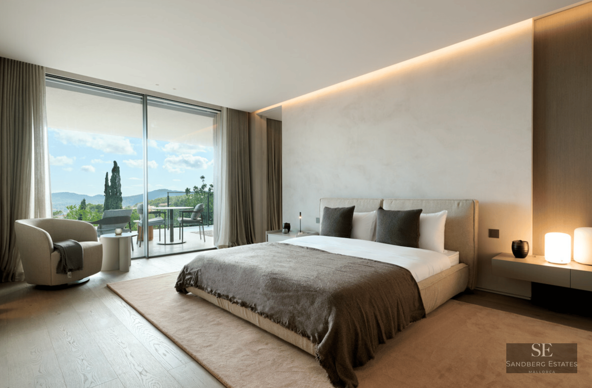 Modern master bedroom with a large bed, designer armchair, and floor-to-ceiling glass doors opening to a scenic terrace.