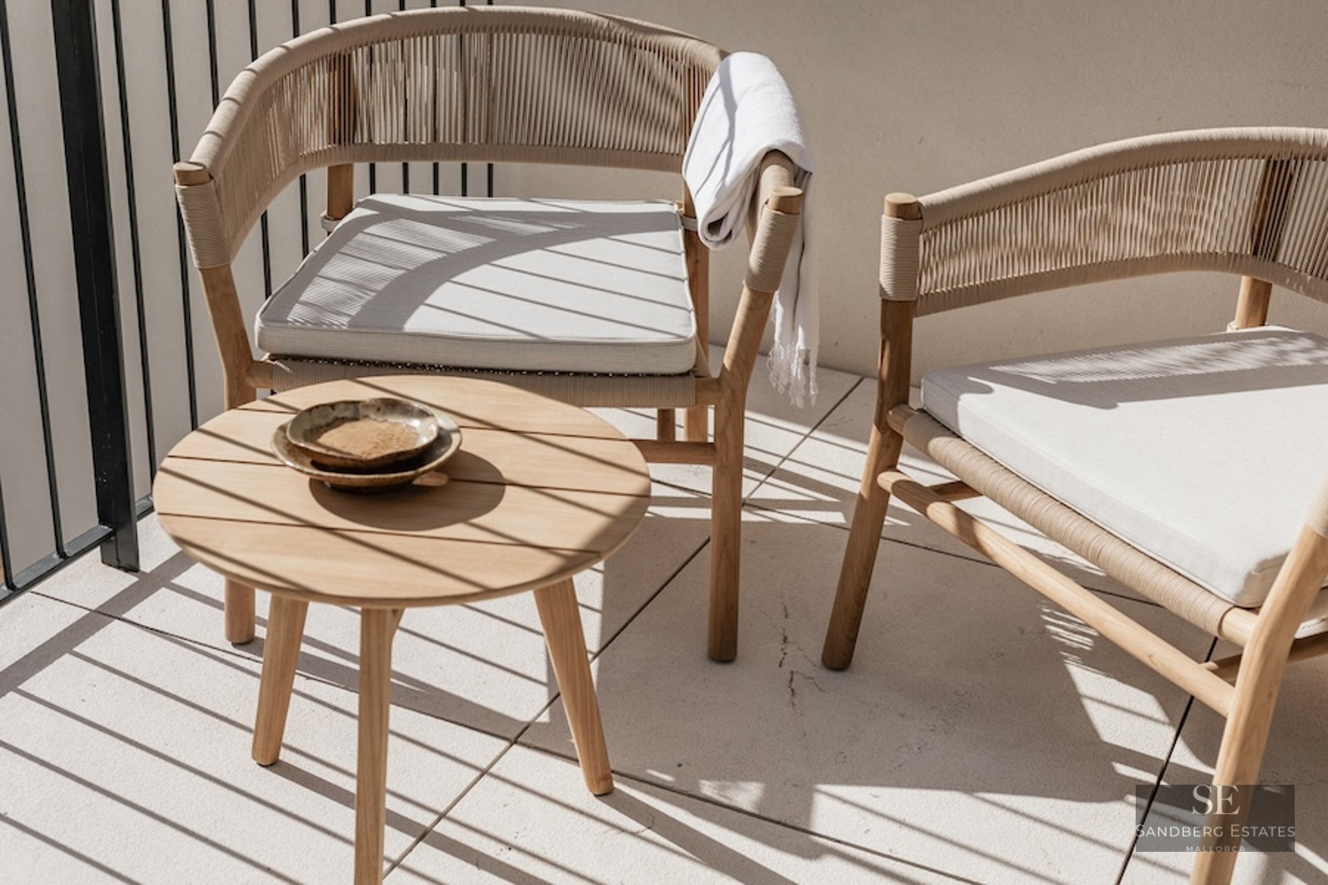 Two elegant woven chairs and a round wooden side table on a sunlit balcony with shadows.