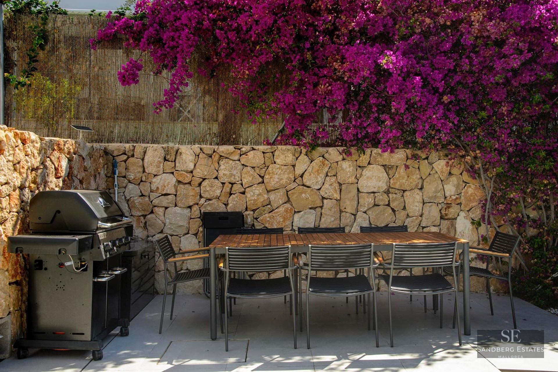 Outdoor patio featuring a wooden dining table, black chairs, gas grill, and a stone wall with vibrant pink flowers.