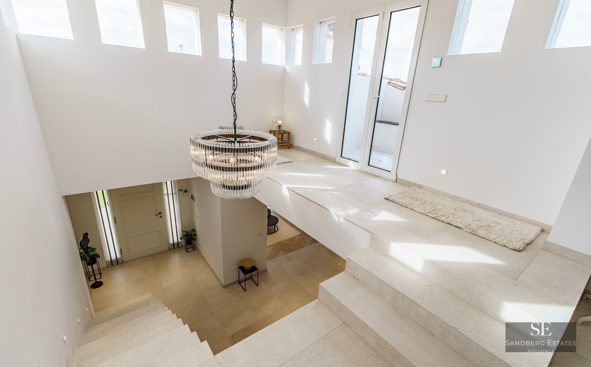 High-angle view of a bright modern foyer with stone stairs, white walls, and a large crystal chandelier.