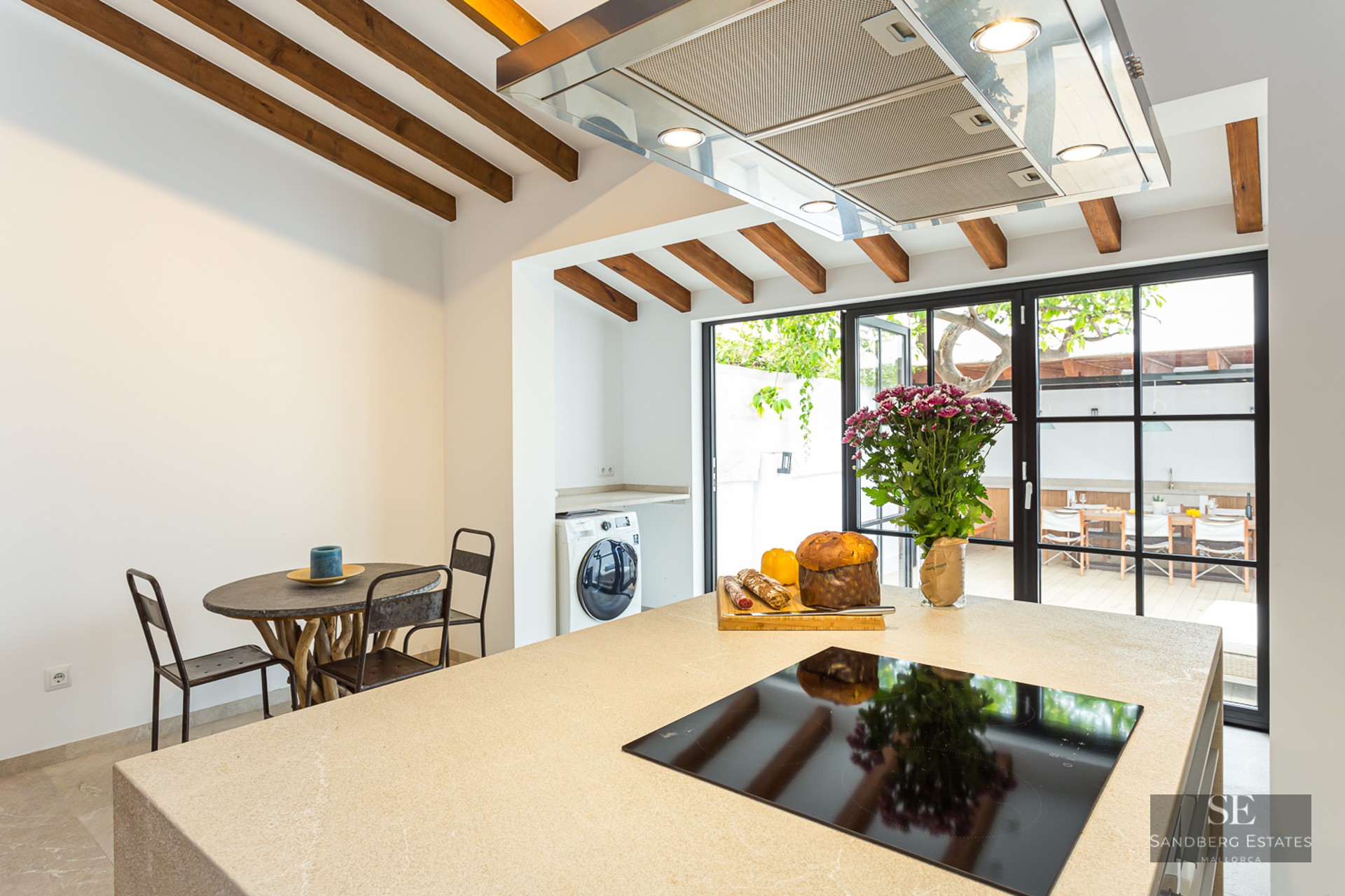 A modern kitchen featuring a beige stone island, induction cooktop, and rustic wooden ceiling beams looking out to a terrace.