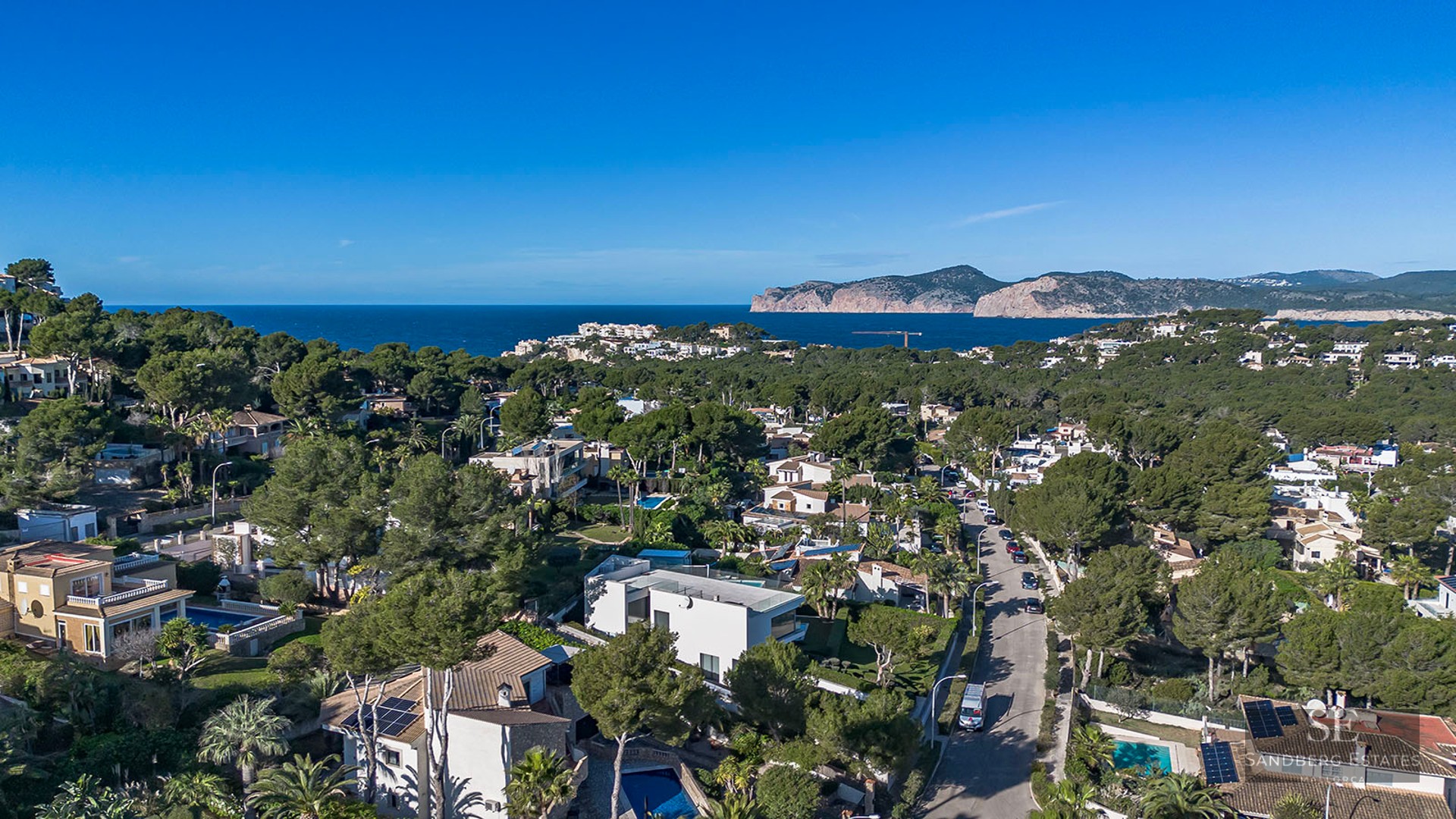 Drone shot of a coastal neighborhood featuring luxury homes, pine trees, and the Mediterranean Sea in the background.