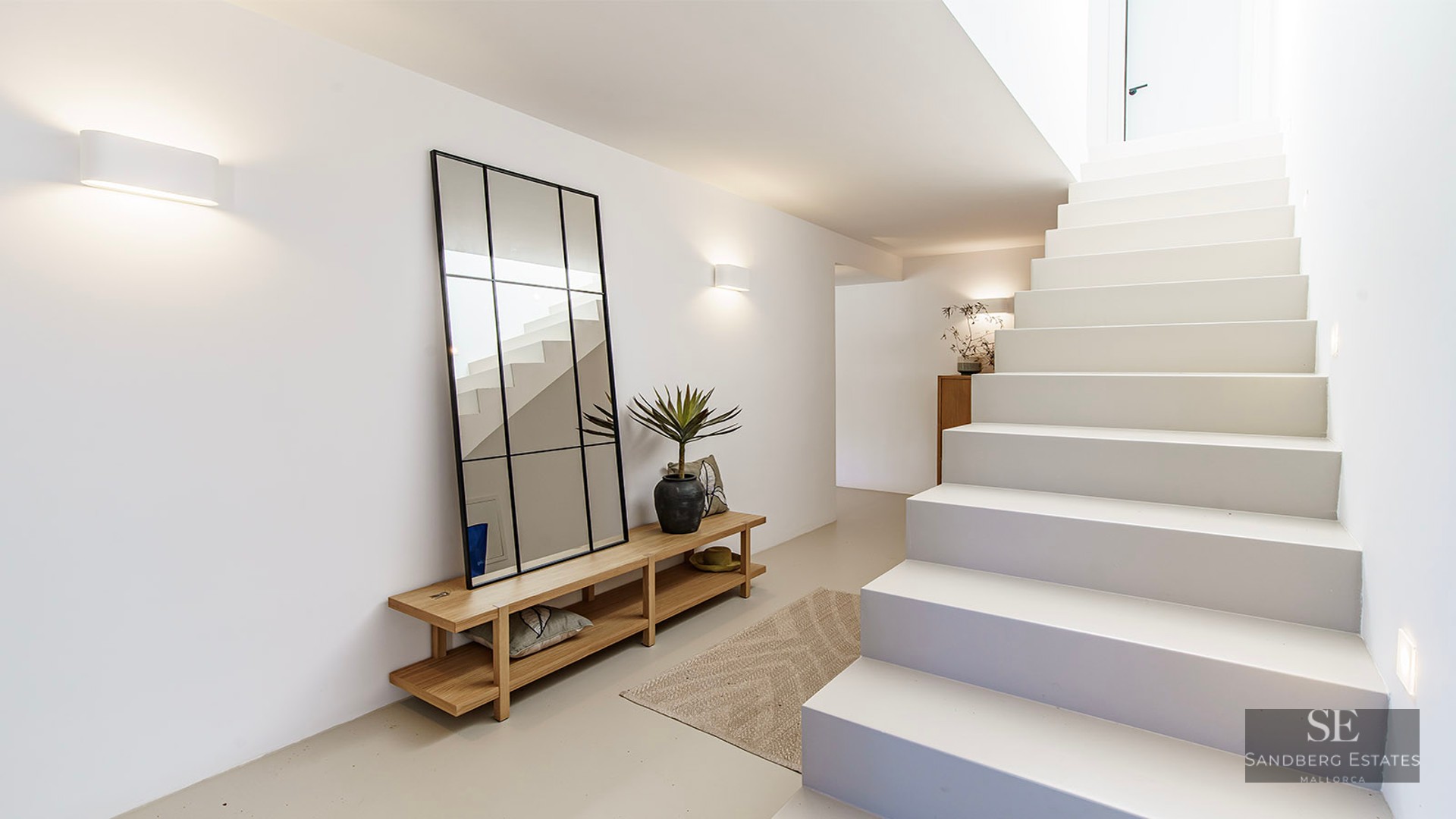 Bright minimalist hallway with white stairs, a large black-framed mirror, wooden bench, and warm wall lighting.