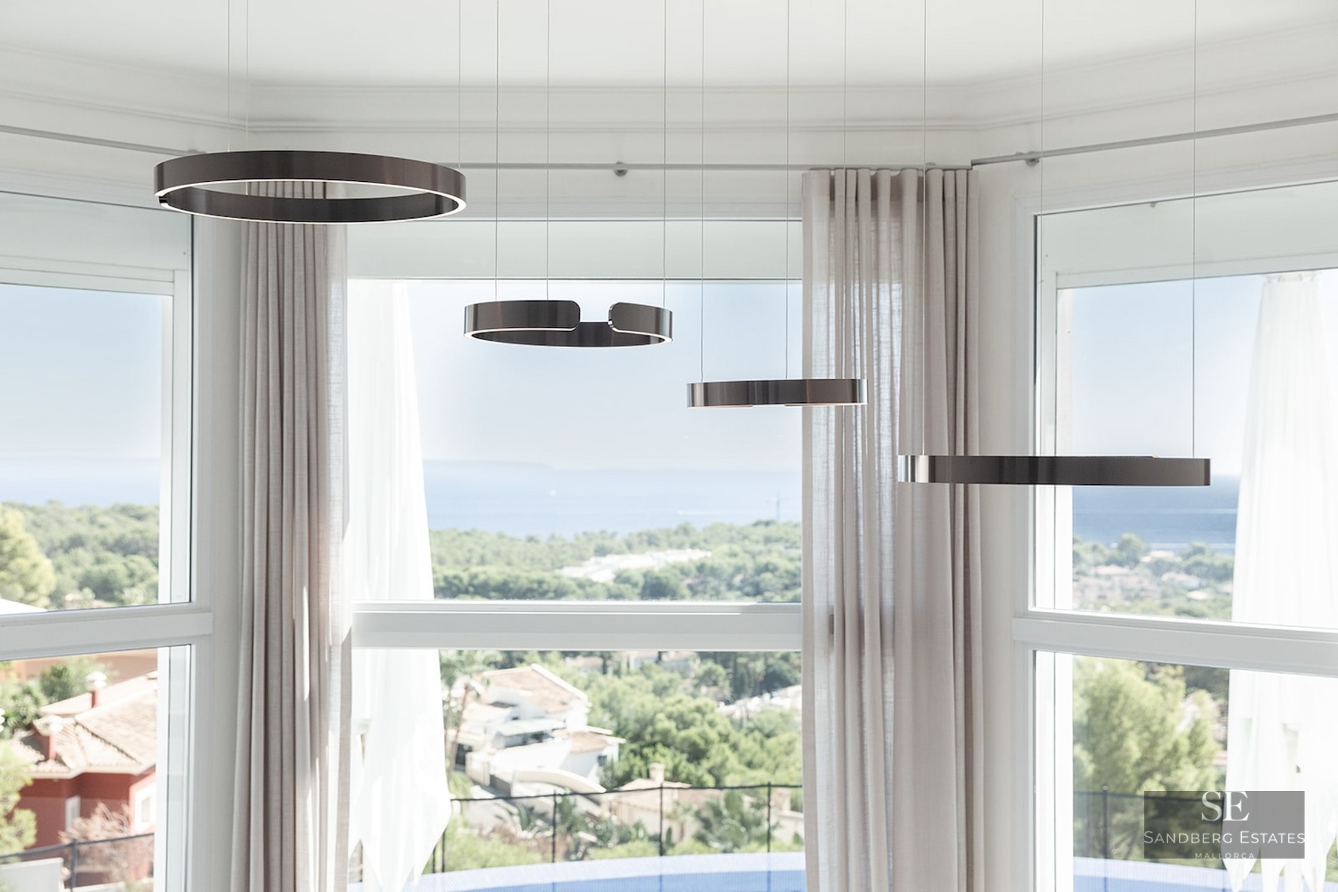Four bronze ring-shaped pendant lights hanging in front of a large bay window overlooking a coastal landscape and the sea.