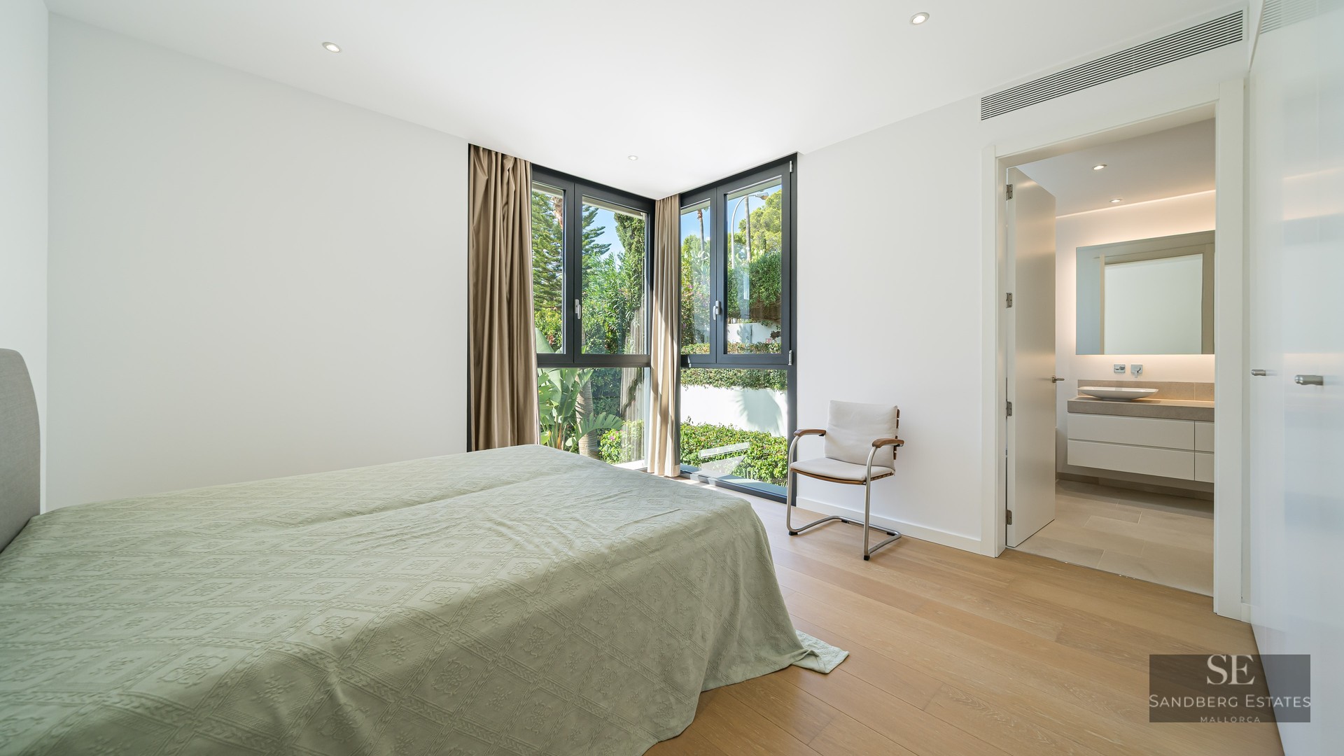 Bright master bedroom with wood floors, large corner windows overlooking greenery, and an adjacent modern bathroom.