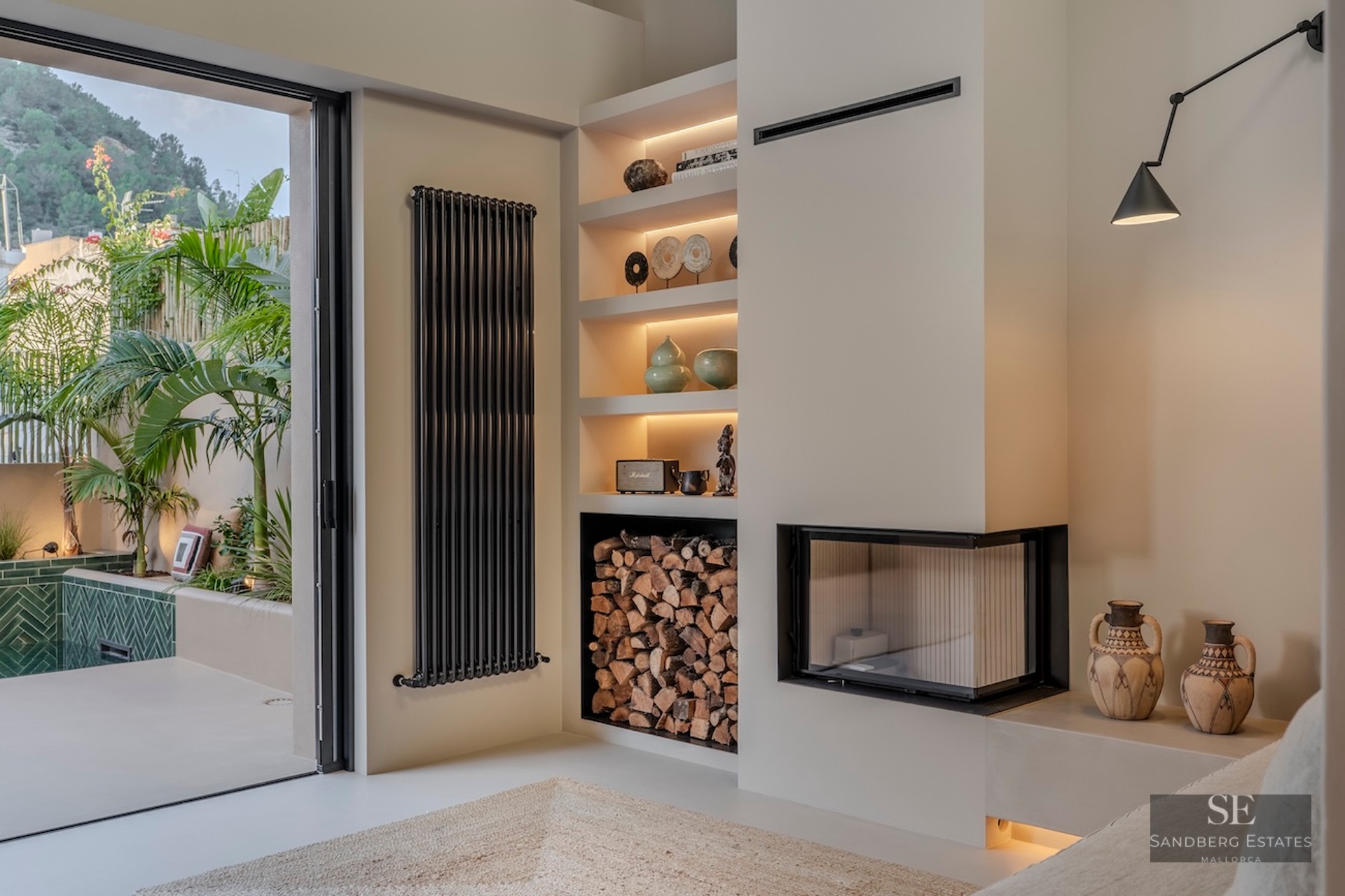 Espace de vie intérieur avec cheminée d'angle moderne, rangement à bois et vue sur une terrasse extérieure luxuriante.