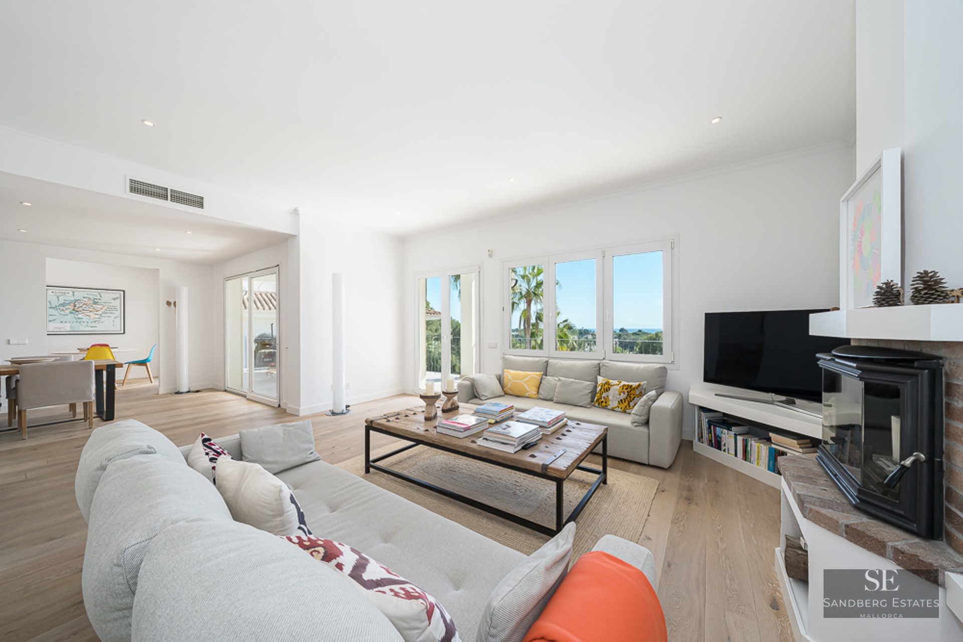Salon lumineux avec canapés gris, table basse en bois, cheminée et grandes fenêtres donnant sur la verdure.