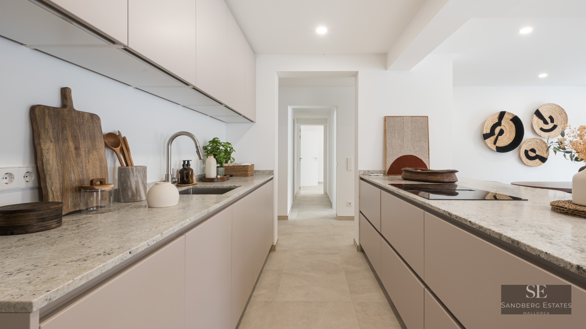 Modern kitchen with beige cabinets, stone countertops, and a view down a white hallway.
