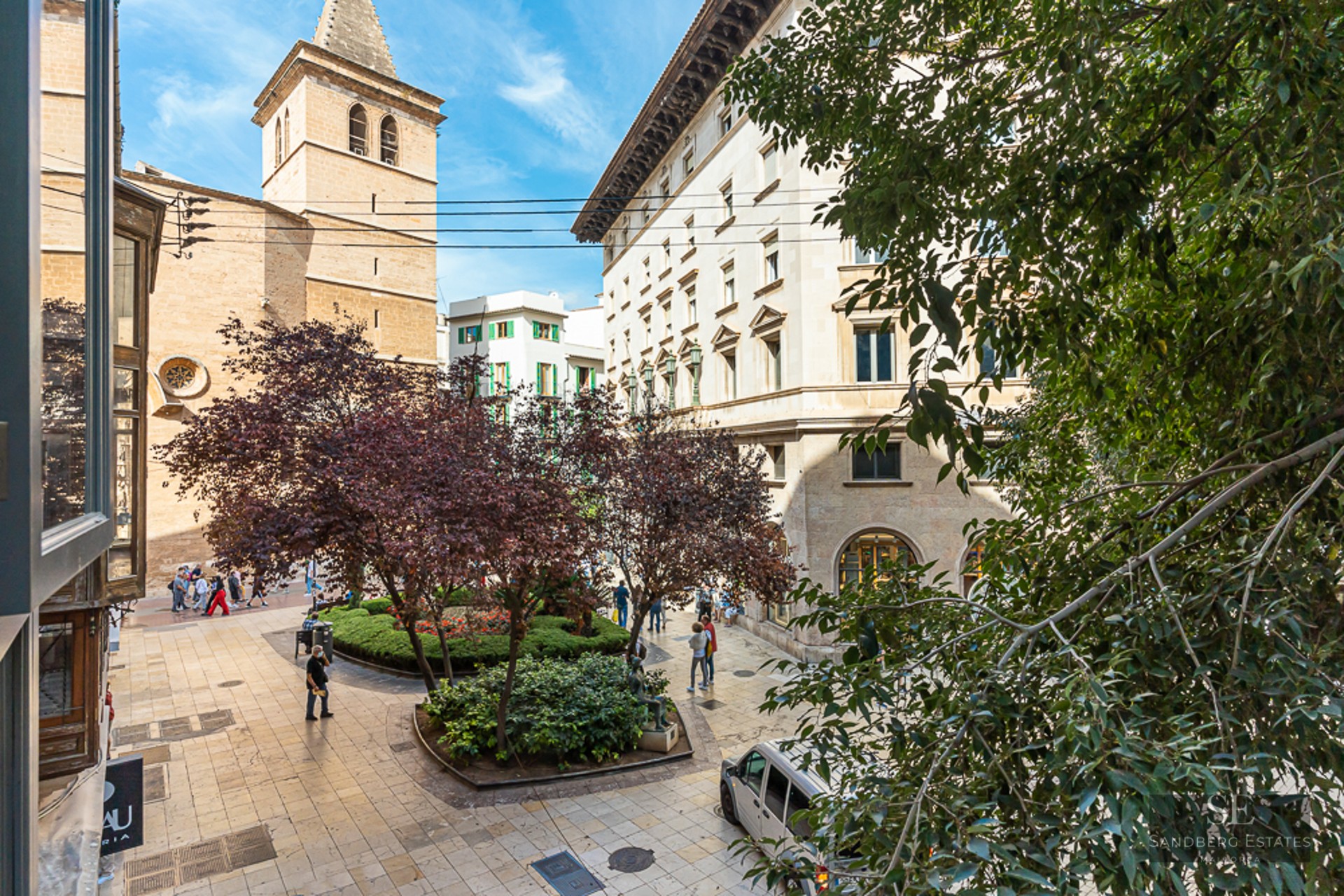 Pedestrian plaza with trees, stone buildings, and a church tower under a clear blue sky.