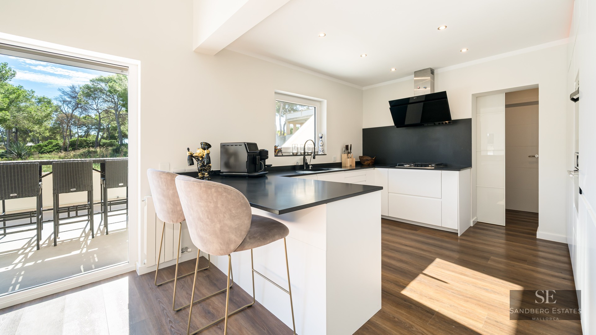 Cuisine moderne blanche avec plans de travail noirs, parquet et bar donnant sur une terrasse ensoleillée.