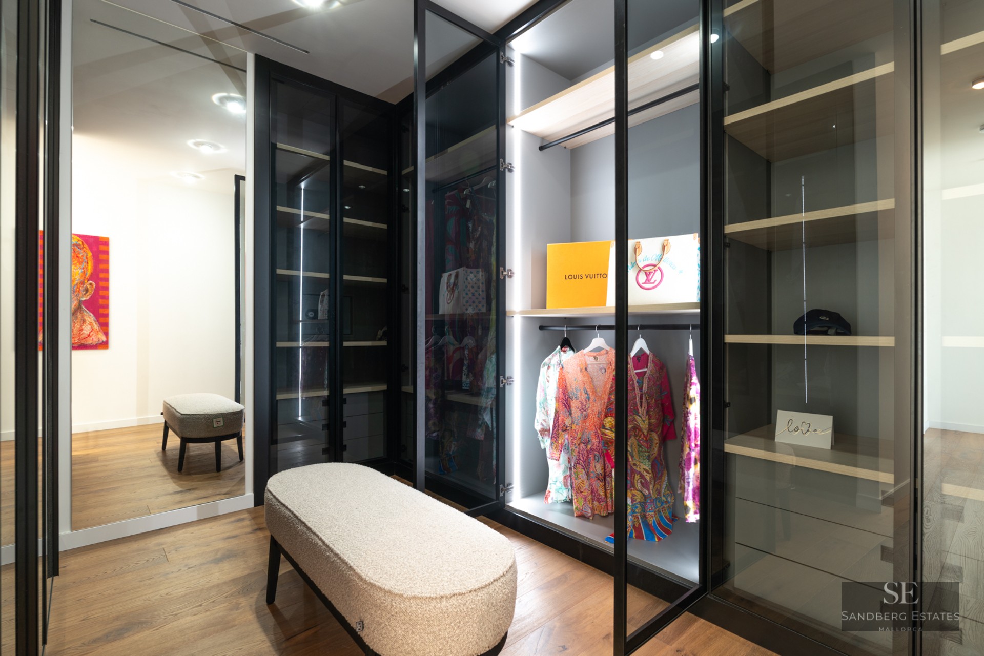 Modern walk-in closet featuring glass-door wardrobes with LED lighting, wood floors, and a textured boucle bench.