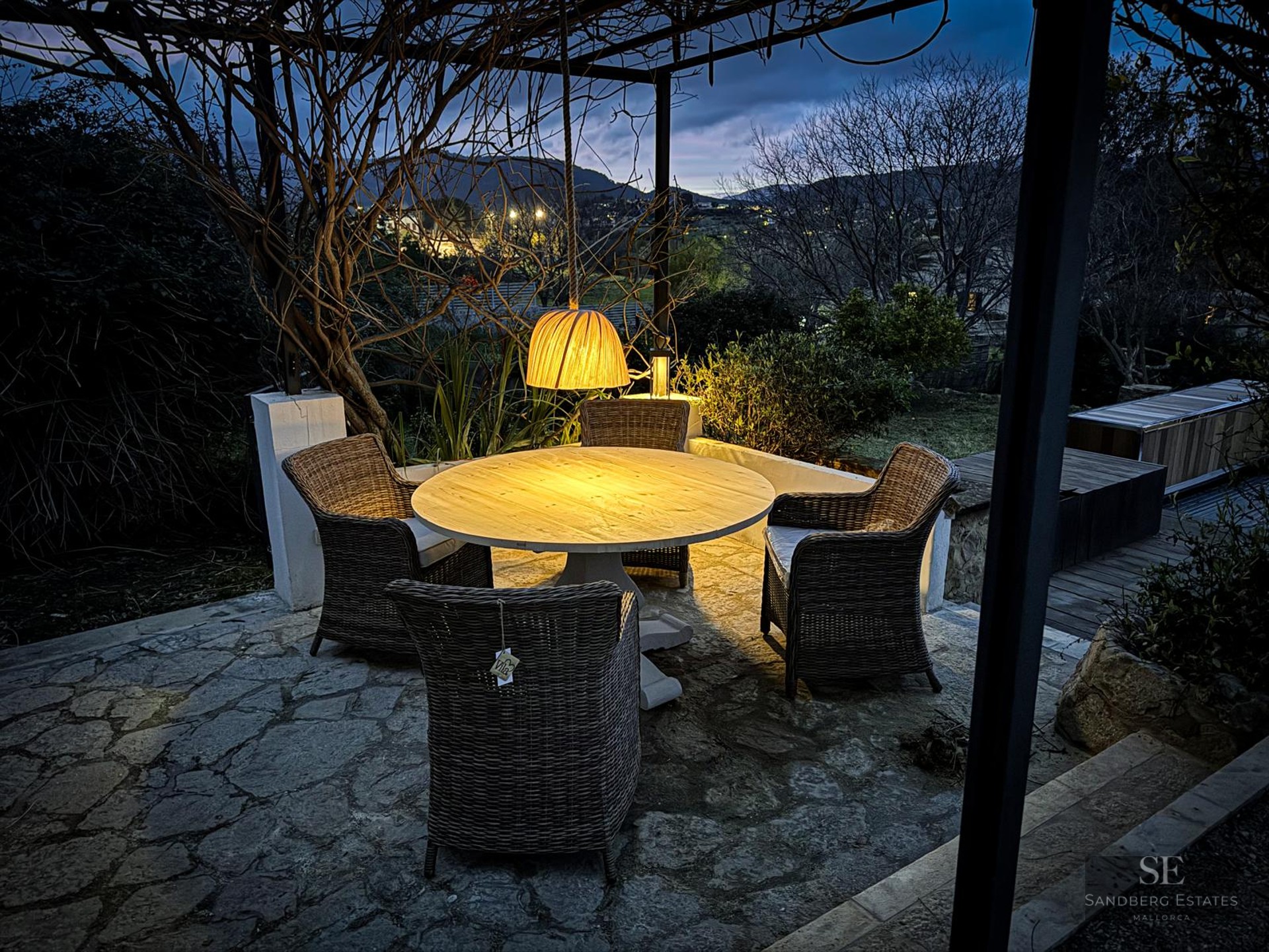 Outdoor stone terrace with a round wooden table, wicker chairs, and a warm pendant light overlooking hills at dusk.