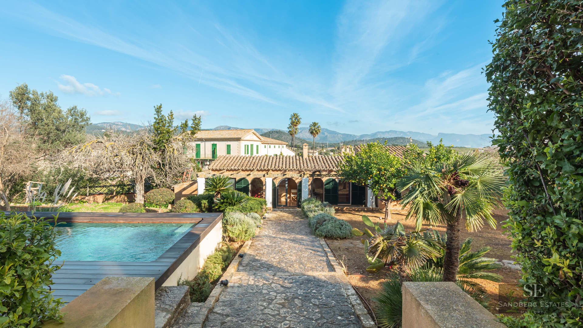 Outdoor swimming pool and stone path leading to a Mediterranean villa with mountain views under a blue sky.