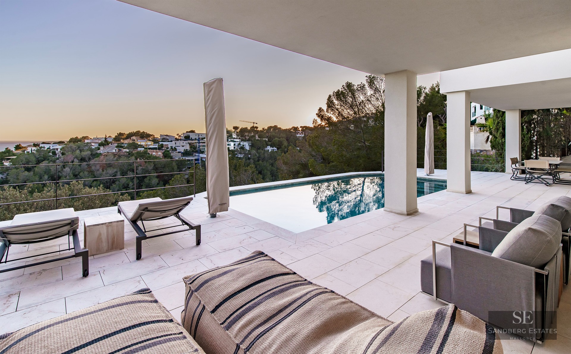 Modern infinity pool on a stone terrace overlooking a lush valley and the sea at sunset with luxury lounge furniture.