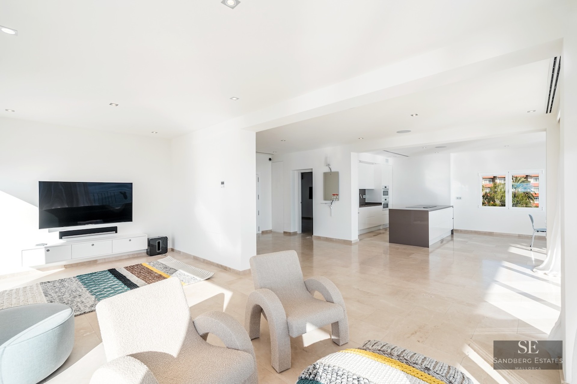Bright, modern living room with plush armchairs, a large wall-mounted TV, and an adjacent open-plan kitchen.