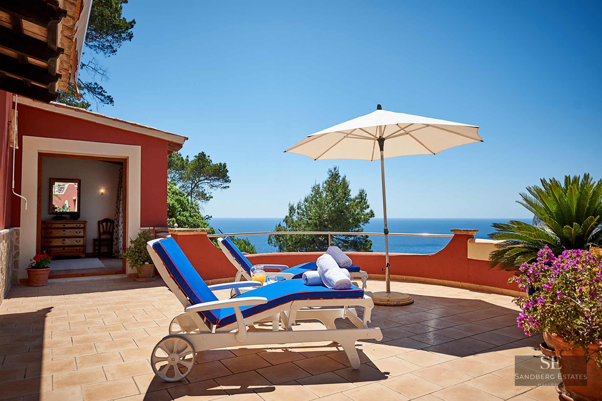 Two blue sun loungers on a terracotta terrace overlooking a bright blue ocean under a clear sky.